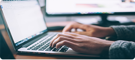 A person's hands over the keyboard of a Macbook. There is a monitor out of focus in the background.