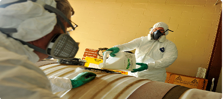 Two men dressed in full personal protective equipment, white boiler suits, filtered mask, green nitrile gloves and safety goggles, filling a large container with bottles of hazardous chemical