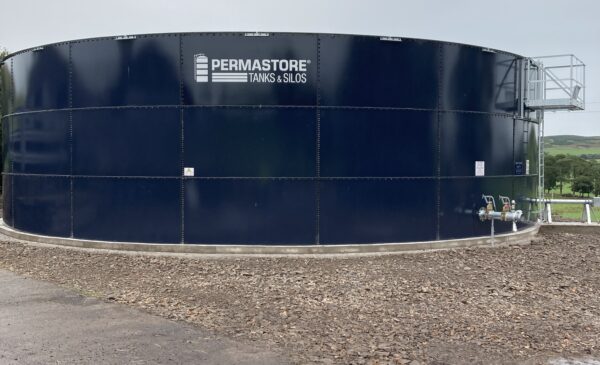Black slurry tank with grey roof on yard behind a hedge. Grass field behind tank.