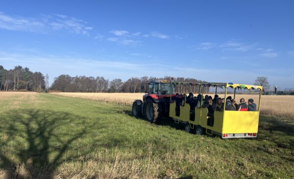 Yellow cart attached to tractor with seats transporting farmers to different locations on farm. Cart and tractor sitting on grass field with farmers inside.
